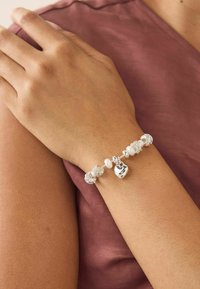 Srebrna bransoletka z zawieszką, zdobiona koralikami, perłami i zawieszką w kształcie serca, noszona na dłoni na mauve tle.