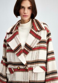 Dobbeltbrystet ternet frakke i crème, brun og rød, med store revers, to forlommer og trækknapper. Blødt, struktureret stof.