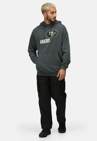 Sudadera de color gris carbón con estampado de "49ERS", bolsillo canguro y puños acanalados. Combinada con pantalones cargo negros y zapatos negros con cordones.