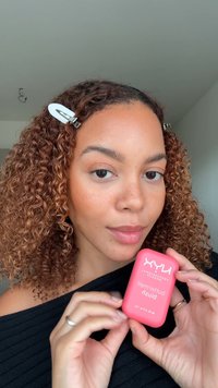 NYX blush product vastgehouden in handen, ovale roze verpakking met witte tekst. Krullend bruin haar, zwarte top, natuurlijke huidskleur met subtiele sproeten.