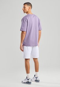 Violetti puuvillapaidat, joissa on lyhyet hihat, yhdistetty valkoisiin shortseihin. Mallilla on päällään valkoiset ja laivastonsiniset tennarit. Tausta on neutraalin harmaa.