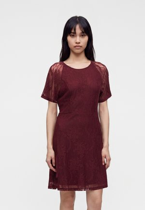 Jeune femme aux longs cheveux foncés portant une robe en dentelle bordeaux à manches courtes, debout devant un fond clair uni.