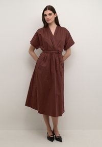Brown wrap dress with short sleeves, a tie waist, and a midi length. Smooth texture and a simple, clean design. Black pointed shoes.