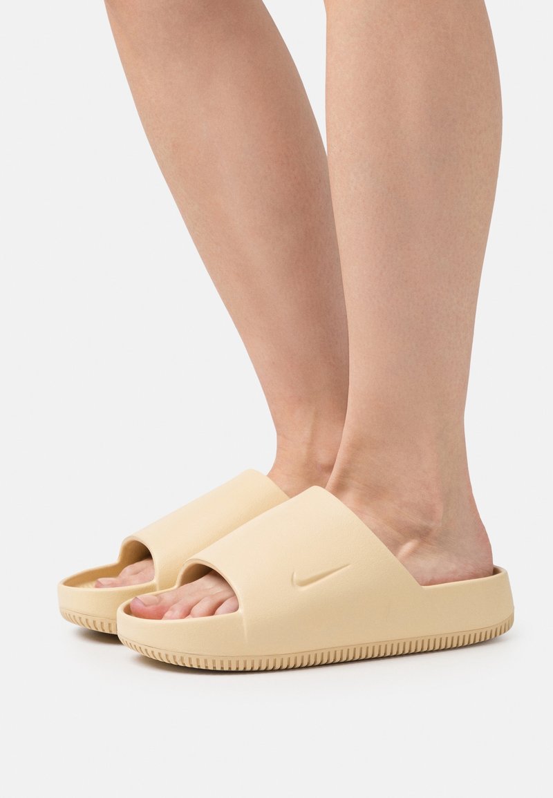 Pieds portant des sandales Nike slide beiges avec une large bride et une semelle texturée sur un fond neutre.