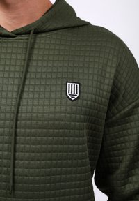 Sudadera verde oliva con un patrón texturizado y acolchado, que presenta un distintivo bordado en blanco y negro en el pecho y una capucha con cordón.