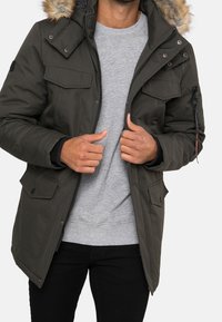 Chaqueta de invierno verde oscuro con capucha forrada de pelo, que cuenta con múltiples bolsillos frontales y un cierre de cremallera. Lleva puesta sobre un suéter gris y pantalones negros.