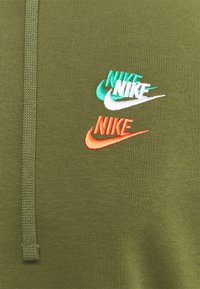 Olivgrön sweatshirt med tre broderade Nike-logotyper i turkos, vitt och orange, samt texturerade dragkedjedetaljer.
