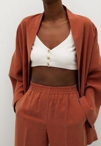 Rdzawy, oversize blazer na białej, krótkiej bluzce z guzikami, w połączeniu z koordynującymi spodniami na gumce, wszystko w miękkim materiale.