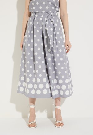 Frau trägt einen grauen Midirock mit weißen Punkten und einem gebundenen Gürtel, kombiniert mit weißen, hochhackigen Riemchensandalen.