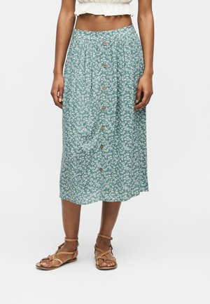 Zielona spódnica midi z białym kwiatowym wzorem, z przodu zapinana na guziki, noszona z beżowymi sandałami na paski i białym krótkim topem