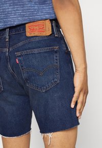 Mörkblå denimshorts med fransade kanter, som har två bakfickor och en läderetikett med röd text på midjebandet.