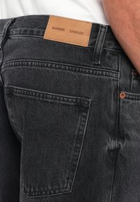 Jean gris foncé avec une étiquette en cuir portant le mot "SAMSE" en relief. Le tissu a une finition texturée avec des coutures visibles et une légère usure.