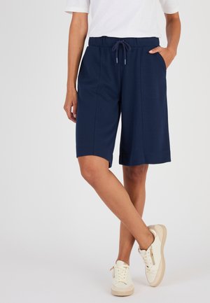 Persona che indossa pantaloncini blu navy alla moda fino al ginocchio con coulisse in vita, maglietta bianca e scarpe da ginnastica bianche, in piedi con una gamba incrociata sull'altra.