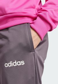 Sudadera rosa con puños acanalados combinada con pantalones jogger grises que presentan un logo blanco de "adidas", textura suave y bolsillos laterales.