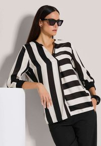 Zwart-wit gestreepte blouse met een kraag, doorzichtige stof en met manchet-mouwen, gecombineerd met zwarte broek en een grote zwarte armband.