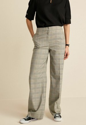 Persoon die beige en zwart geruite wijd uitlopende broek draagt, zwarte korte mouwen top, zwart horloge en zwarte sneakers met witte zolen.