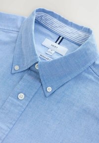 Chemise bleu clair à col en tissu texturé, avec des boutons blancs et un col intérieur rayé. L'étiquette indique "NEXT" taille M.