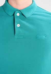 Polo shirt turchese in tessuto testurizzato, con patta per i bottoni a due, colletto e logo impresso sul petto.