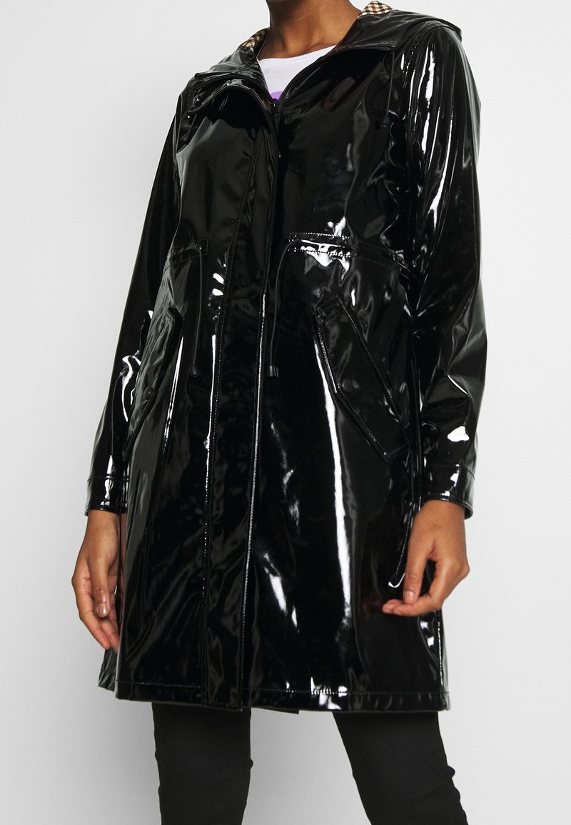 Person trägt eine glänzend schwarze Kapuzenregenjacke mit Fronttaschen und Reißverschluss, teilweise geöffnet, um ein weißes Hemd darunter sichtbar zu machen.