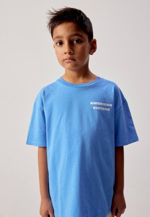 Fant z kratkimi lasmi, oblečen v ohlapno modro majico s tekstom "AMERICAN VINTAGE", stoji pred enostavnim svetlim ozadjem.