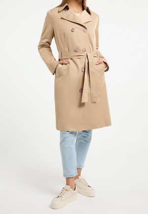 Vrouw draagt een beige trenchcoat met dubbele rij knopen en een tailleceintuur, lichtblauwe cropped jeans en beige plateau sneakers.