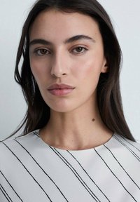 Bílý top s černými vertikálními pruhy, hladká textura, kulatý výstřih. Jemný, přirozený make-up, dlouhé tmavé vlasy, neutrální pozadí.