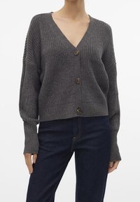 Harmaa neulottu neuletakki, jossa on syvä V-pääntie, pitkät hihat ja kolme etunappia. Kankaan tekstuuri ja lyhyt siluetti.