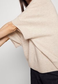 Beiger Strickpullover mit weiten Ärmeln, gerippten Säumen und weicher Textur, kombiniert mit dunklen Hosen. Der Pullover hat einen lockeren, entspannten Schnitt.