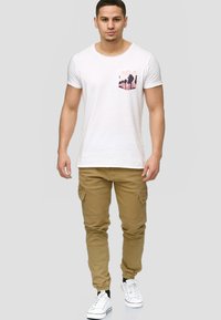 Fehér, rövid ujjú póló, mellzsebbel, rózsaszín pálmafás grafikával, bézs cargo nadrággal és fehér sportcipővel párosítva.