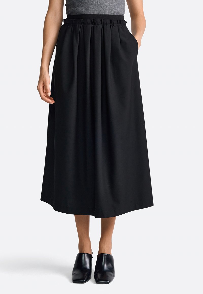 Minifalda negra con pliegues y banda elástica en la cintura, con bolsillos laterales y un diseño fluido. Combinada con mules negros sobre un fondo claro.