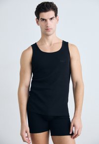 Schwarzes Tanktop aus glattem Stoff mit breiten Trägern, das ein dezentes Logo trägt. Trägt man mit passenden schwarzen Shorts, die beide eng anliegen und schlank im Design sind.