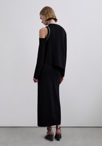 Mujer con una blusa negra de manga larga con hombros descubiertos y falda larga a juego, llevando sandalias negras de tacón, de pie contra una pared lisa.