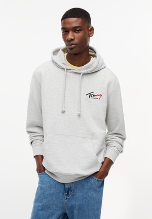 Młody mężczyzna z rękami w kieszeniach, ubrany w jasnoszary kaptur Tommy Jeans i niebieskie dżinsy, stojący na tle jednolitego białego tła.