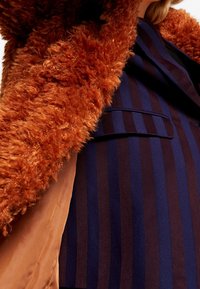 Brun imiteret pelsjakke med en fluffy tekstur, kombineret med et mørkeblåt og vinrødt stof med nålestriber under; har en enkelt lomme.