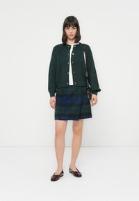 Groene gebreide cardigan, witte top en geruiten rok in blauwe en groene tinten. Bruine knopen, zwarte handtas en bordeauxkleurige platte schoenen.