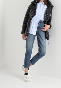 Černá péřová bunda s lesklým povrchem, světle modrá košile na knoflíky, potrhané modré džíny a bílé tenisky s tmavými akcenty.