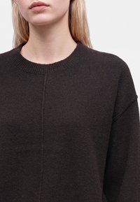 Jeune personne à la peau claire et aux longs cheveux blonds portant un pull en tricot noir avec un col rond côtelé.