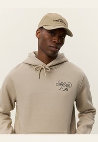 Sudadera beige con un bolsillo frontal y cordón, con el logo bordado en negro "Les Deux NY, NY". Gorra beige a juego con texto.