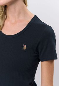 Tummansininen ribbineulottu t-paita, jossa pyöreä pääntie ja lyhyet hihat. Rinnan kohdalla pienellä kirjotulla logolla kontrastivärein.