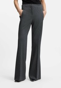 Donna che indossa pantaloni a gamba larga grigio scuro a vita alta con le mani nelle tasche e scarpe nere, in piedi su uno sfondo bianco semplice.
