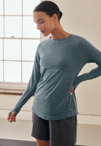 Långärmad träningsskjorta i teal, tillverkad av strukturerad väv, med rund halsringning och tumhål. Kombinerad med mörka shorts.