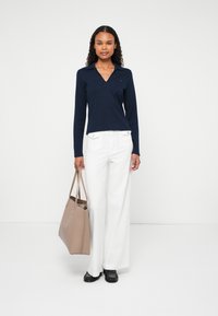 Marineblå langermet V-hals topp kombinert med hvite høytlivsbukser. Modellen holder en beige tote veske og har på seg svarte sko. Enkel design og farger.