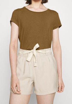 Kobieta w brązowej bluzce z krótkim rękawem, wpuszczonej do beżowych szortów z wysokim stanem, z materiałowym paskiem zawiązanym w kokardę w talii.