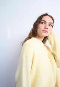 Jeune femme aux cheveux ondulés portant un pull en maille jaune doux, regardant légèrement vers le haut et touchant ses cheveux sur un fond clair uni.