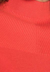 Roter Strickpullover mit Rollkragen, der über eine gerippte Struktur und eine figurbetonte Passform verfügt. Nahtdetails schaffen visuelles Interesse auf der Oberfläche.