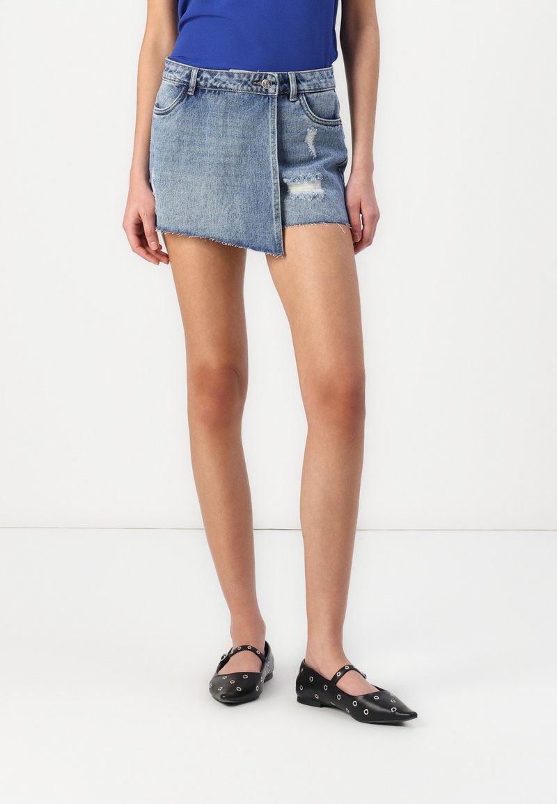 Femme portant une mini-jupe en jean délavé bleu clair asymétrique, un haut bleu et des ballerines noires avec des détails en œillets argentés, debout devant un fond blanc.