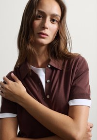 Camisa de polo de punto marrón con cuello y puños blancos, con mangas cortas y detalles de botones, llevada por un modelo con los brazos cruzados.