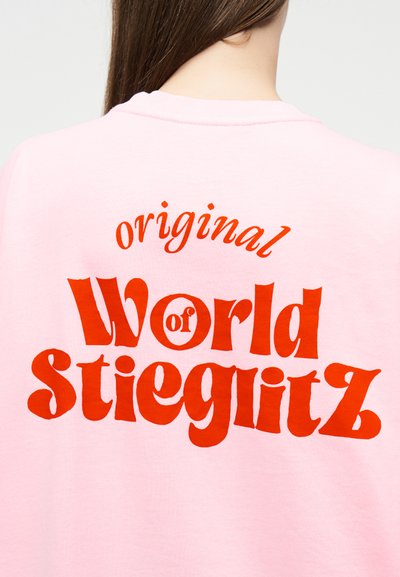 Άτομο με μακριά καστανά μαλλιά, ορατό από πίσω, φοράει ανοιχτό ροζ πουκάμισο με κόκκινο κείμενο "original World of Stieglitz" στην πλάτη.