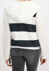 Huvtröja med vit framdel och marinblå ribbad baksida som har horisontella ränder. Avslutad med en praktisk dragsko vid fållen.