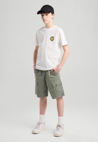 T-shirt bianco in cotone con un logo circolare colorato, pantaloni cargo verde oliva, calze bianche e sneakers beige, abbinati a un cappellino nero.